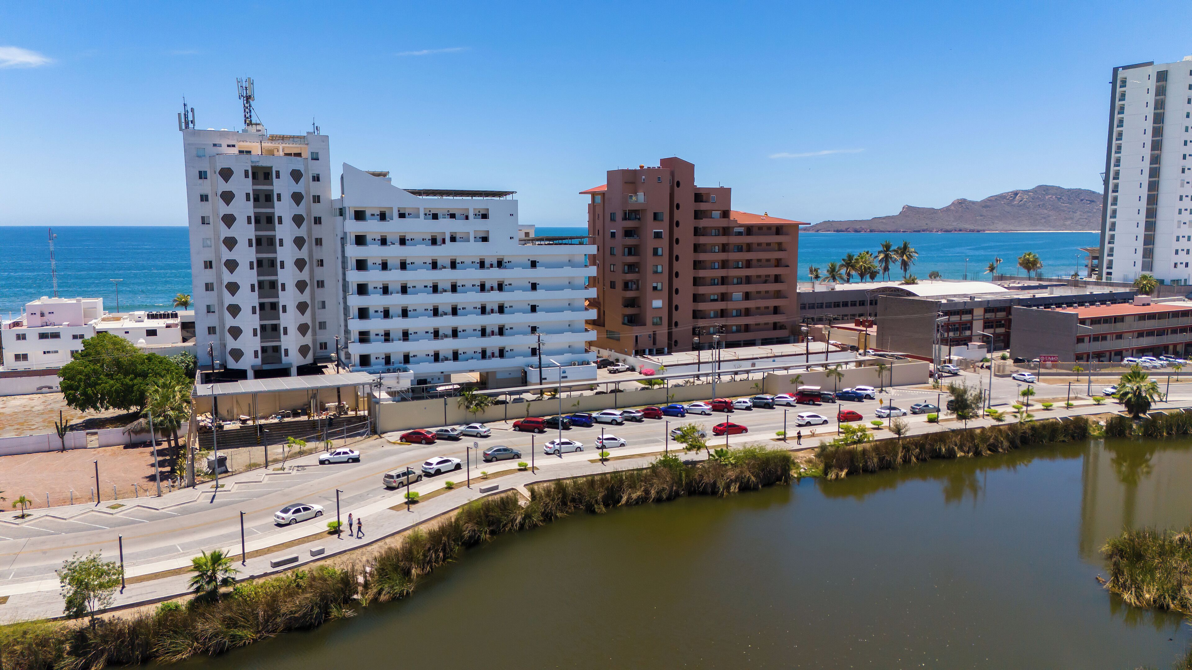 Un paisaje urbano costero con edificios modernos, un puerto deportivo y una avenida con coches estacionados.