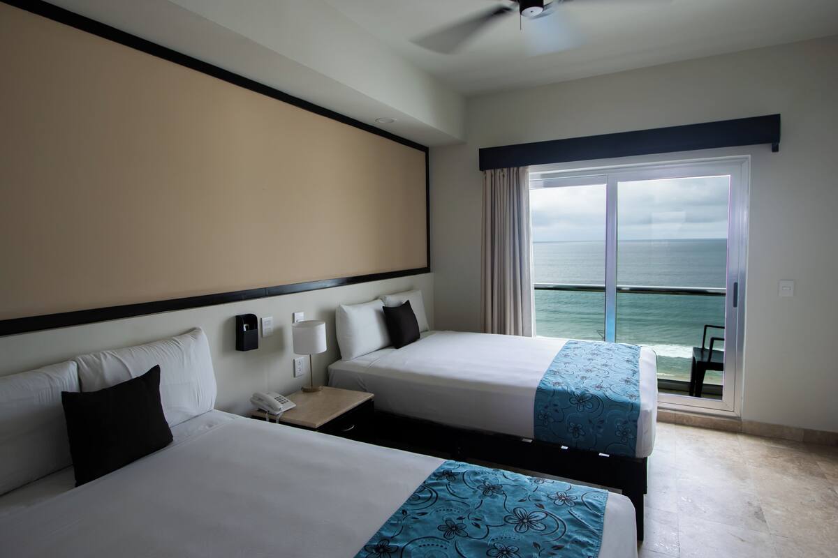 A hotel room with two beds, a large window with a view of the sea, a ceiling fan, and a nightstand with a phone.