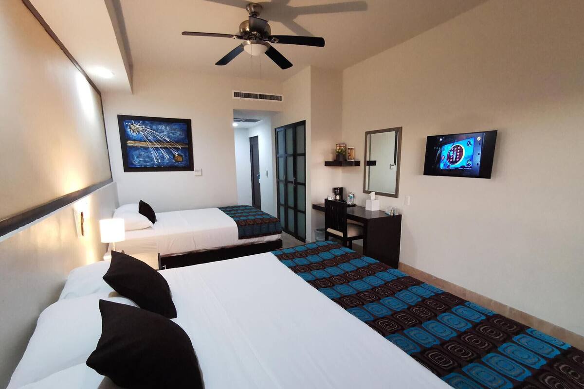 A hotel room with two beds, a desk, a chair, a ceiling fan, a TV, and a door leading to another room.