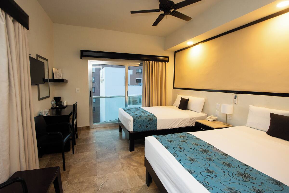 A hotel room with two beds, a desk, a chair, a ceiling fan, and a view of the outside.