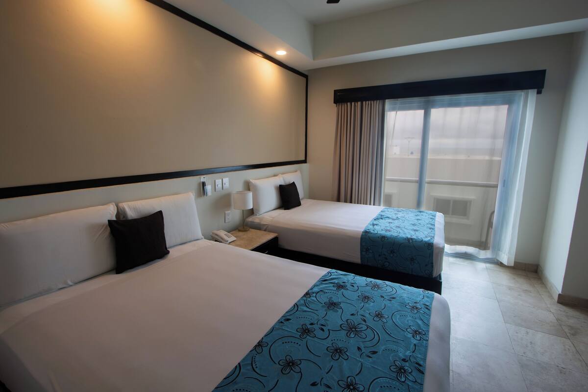 A hotel room with two beds, a large window with curtains, a bedside table with a phone, and a wall-mounted light.