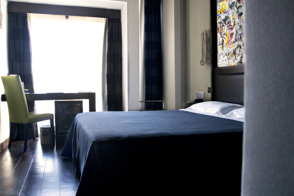 A hotel room with a bed, a chair, a desk, and a window with curtains.