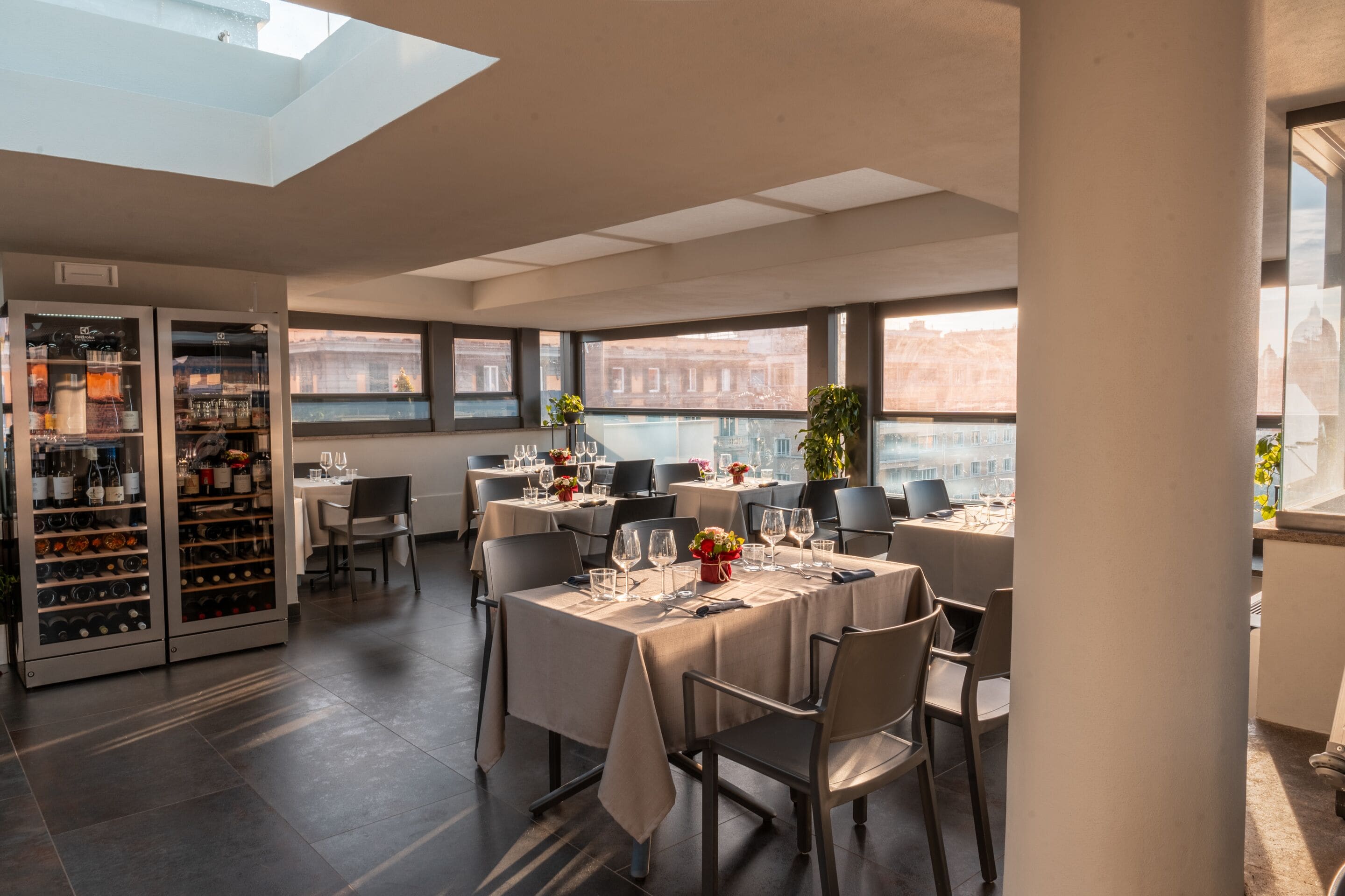 Un comedor moderno con mesas preparadas para dos, un enfriador de vino y grandes ventanales con vistas a la ciudad.