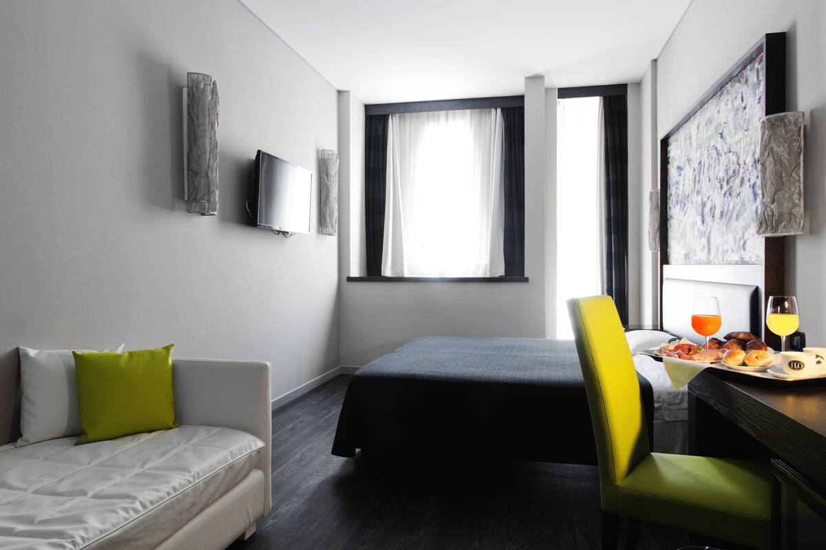 A hotel room with a bed, a sofa, a dining table with breakfast, a TV, and a window with curtains.