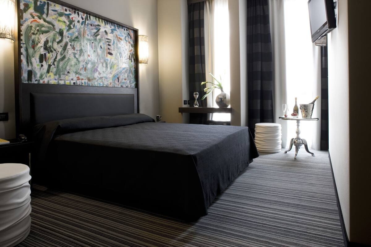 A hotel room with a large bed, a headboard with artwork, a bedside table with a lamp, a small table with a vase and wine glasses, and a view of the outdoors through a window.