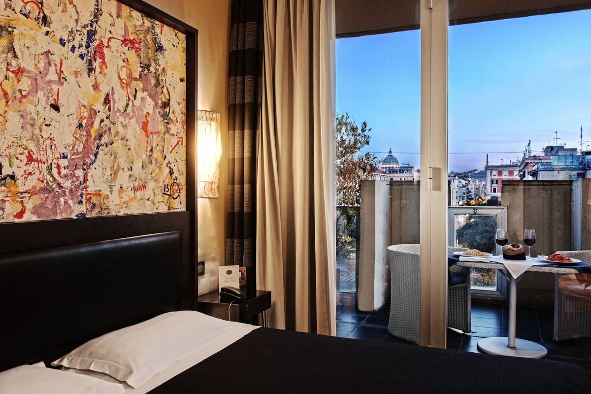 A hotel room with a large bed, a dining area with a table and chairs, and a view of the cityscape through the window.