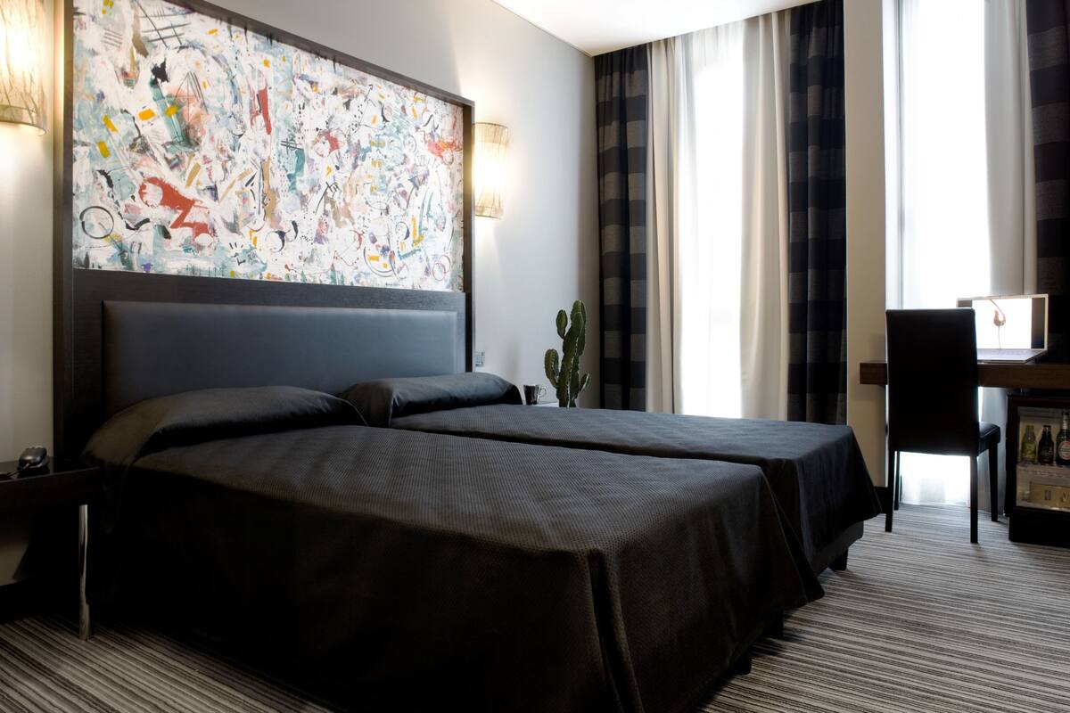 A hotel room with two beds, a large abstract painting on the wall, a cactus on a side table, and a minibar.