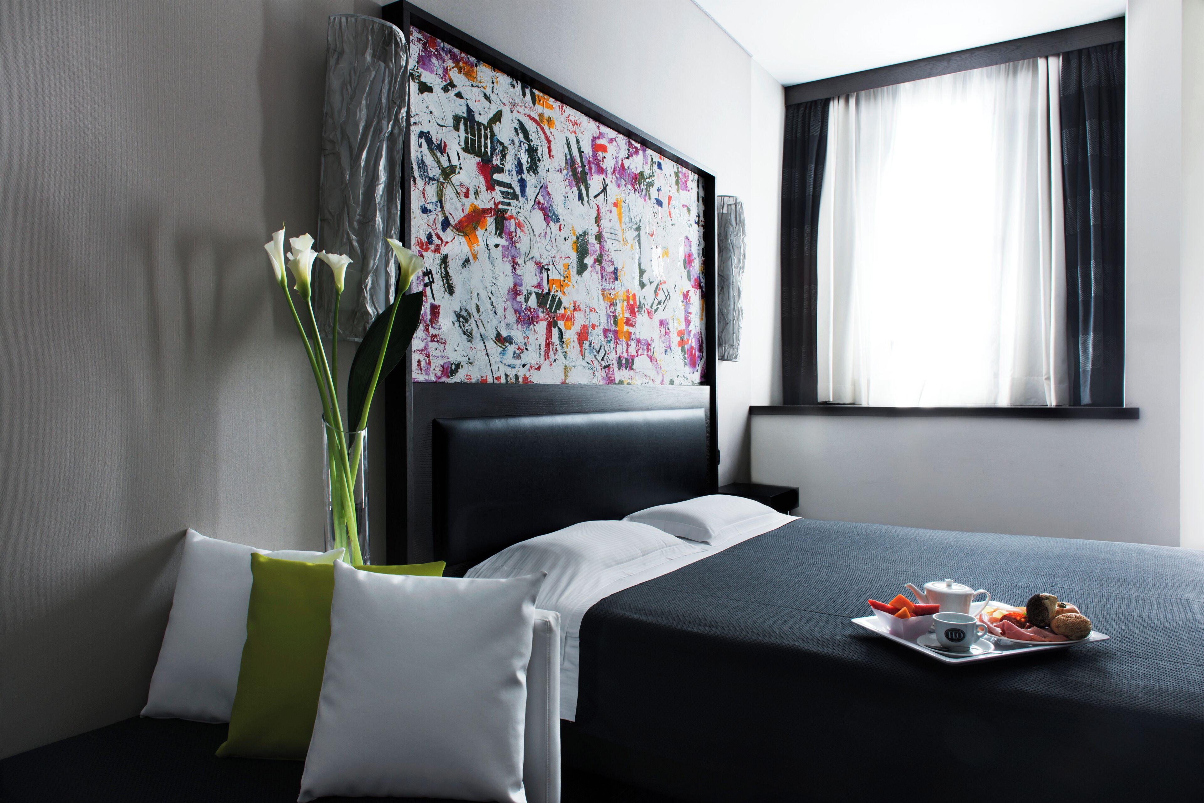Ein Hotelzimmer mit Bett, einem Bild an der Wand, einem Fenster mit Vorhängen und einem Tablett mit Speisen und einer Tasse auf dem Bett.