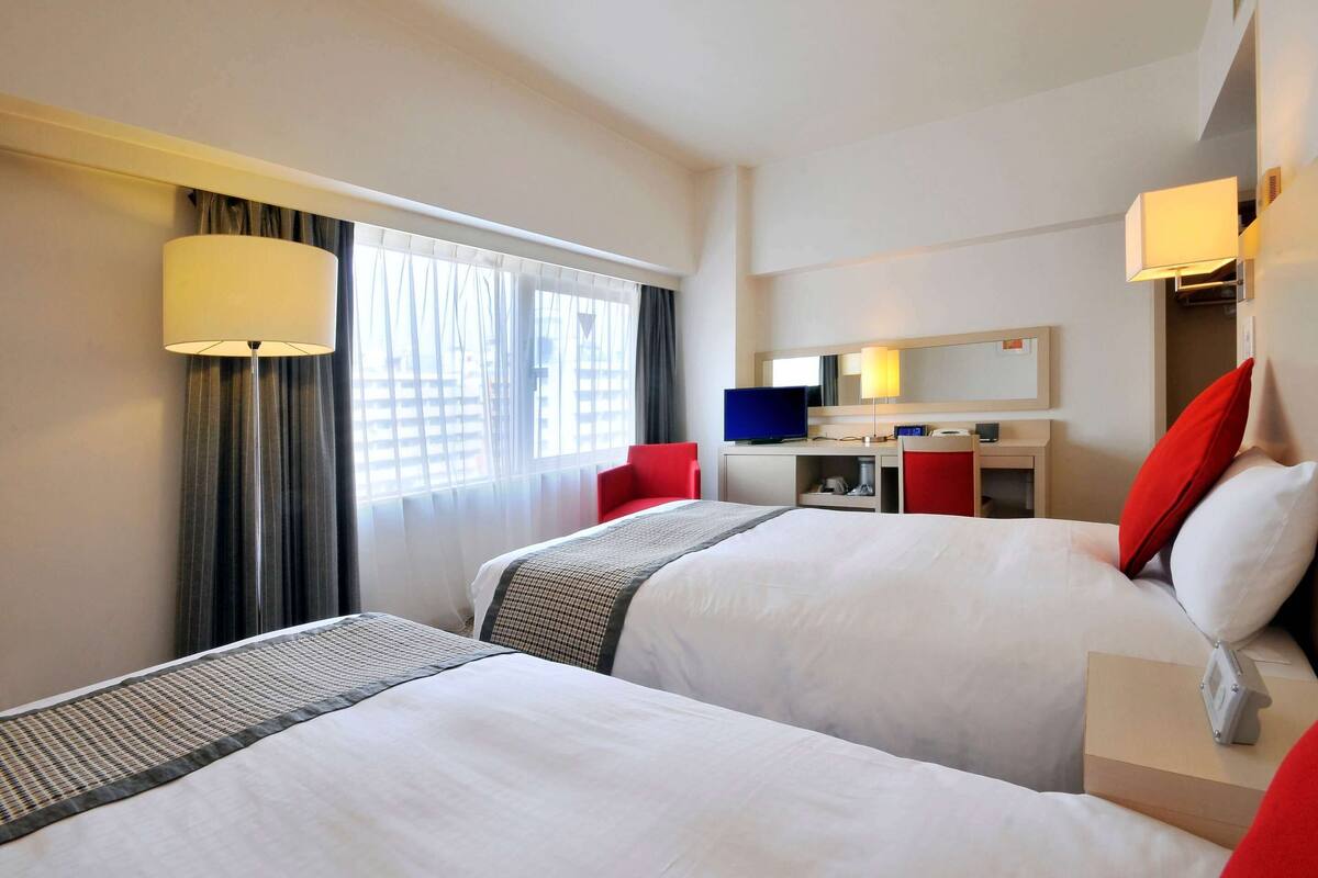 A hotel room with two beds, a desk with a computer, a mirror, and a window with curtains.