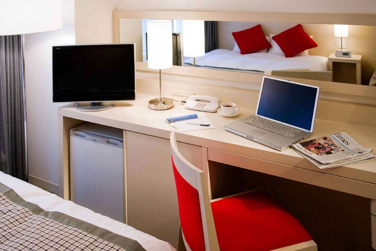 A hotel room with a bed, a desk with a laptop, a television, a telephone, and a newspaper.