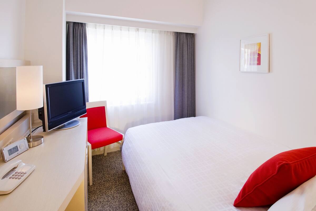 A hotel room with a bed, a desk with a computer monitor, a telephone, and a chair.