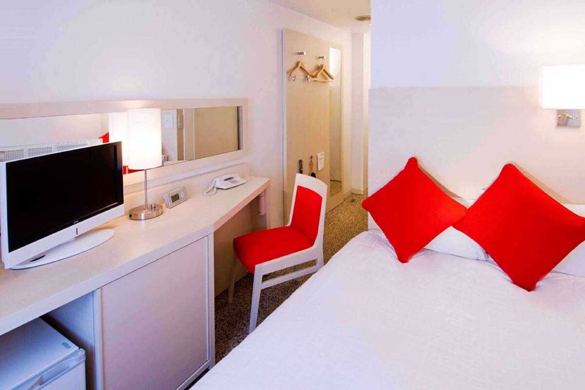 A hotel room with a bed, a desk with a TV, a chair, and a mirror.