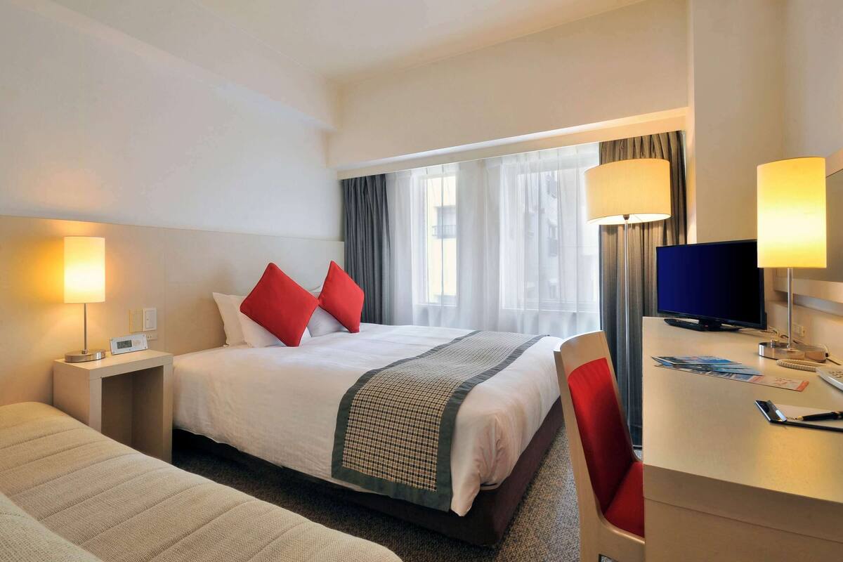 A hotel room with a bed, bedside table with a lamp, a desk with a computer, and a chair.