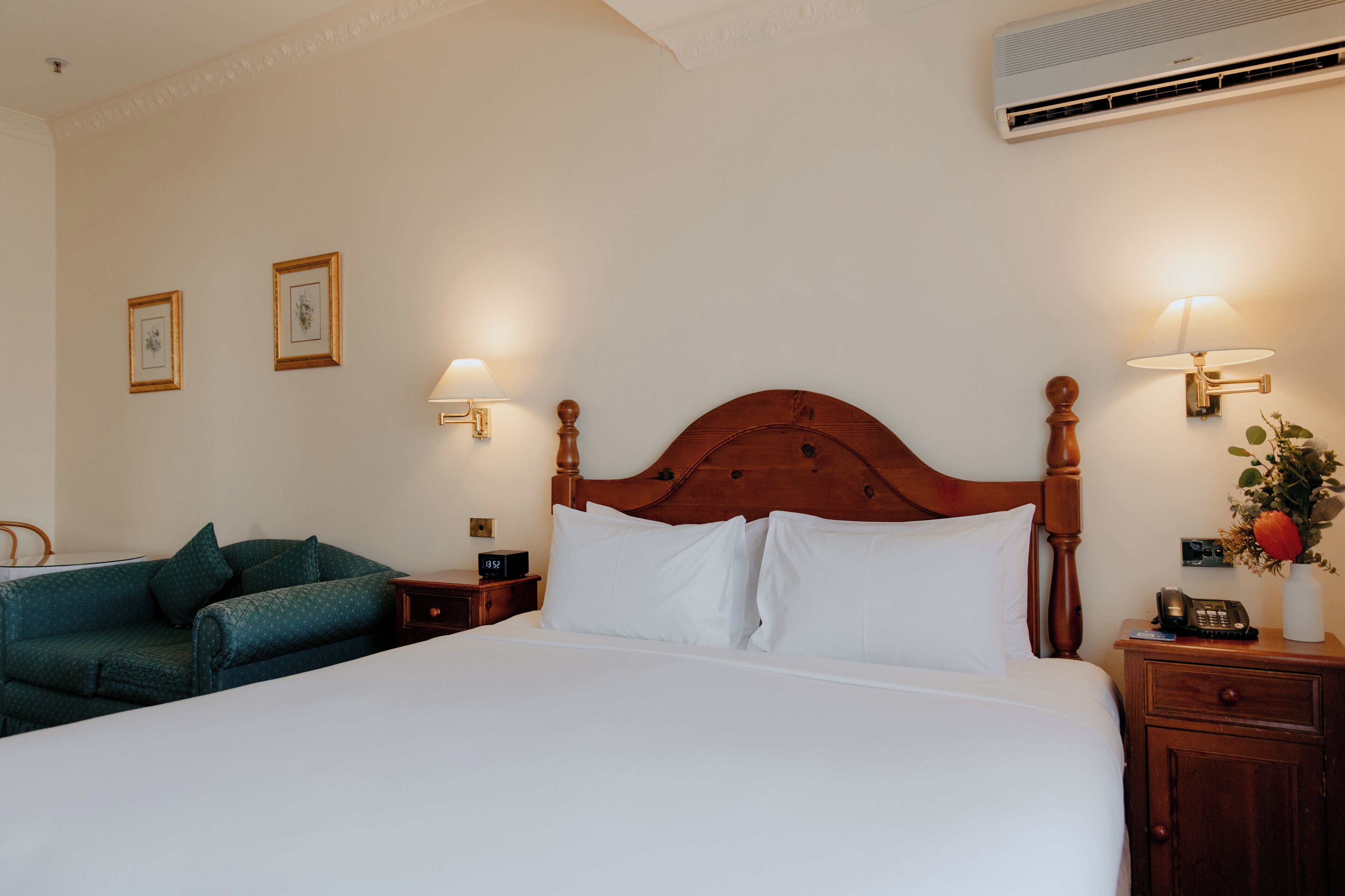 Un lit bien fait, avec du linge de lit blanc, une tête de lit en bois, des lampes de chevet, une table de chevet avec un téléphone et un canapé vert dans une chambre d’hôtel.