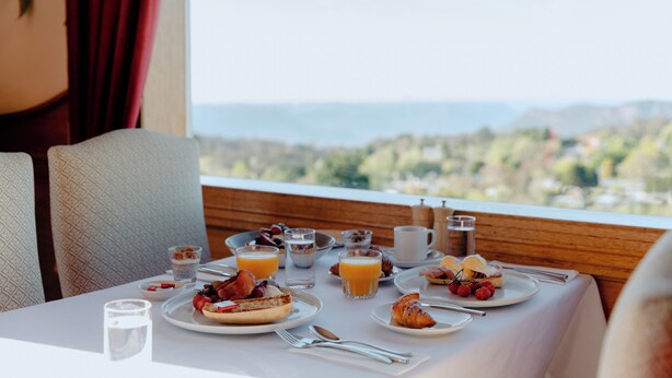 Una mesa de desayuno dispuesta con platos de comida, vasos de jugo de naranja y vistas a un paisaje pintoresco.