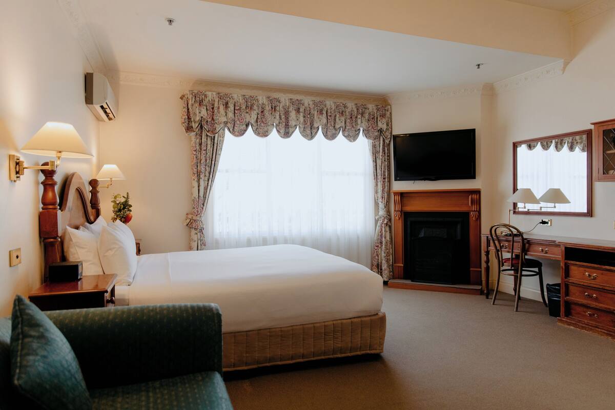 A hotel room with a large bed, a desk with a chair, a television, and a fireplace.