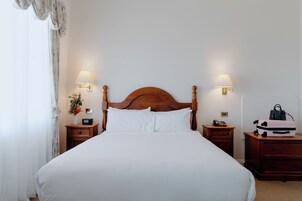 Una cama bien hecha con sábanas blancas, dos mesitas de noche con lámparas y una maleta sobre una de ellas.