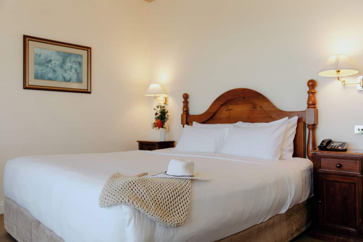 A neatly made bed with white linens, a wooden headboard, and a white hat on the bed. There are two bedside lamps and a framed picture on the wall.