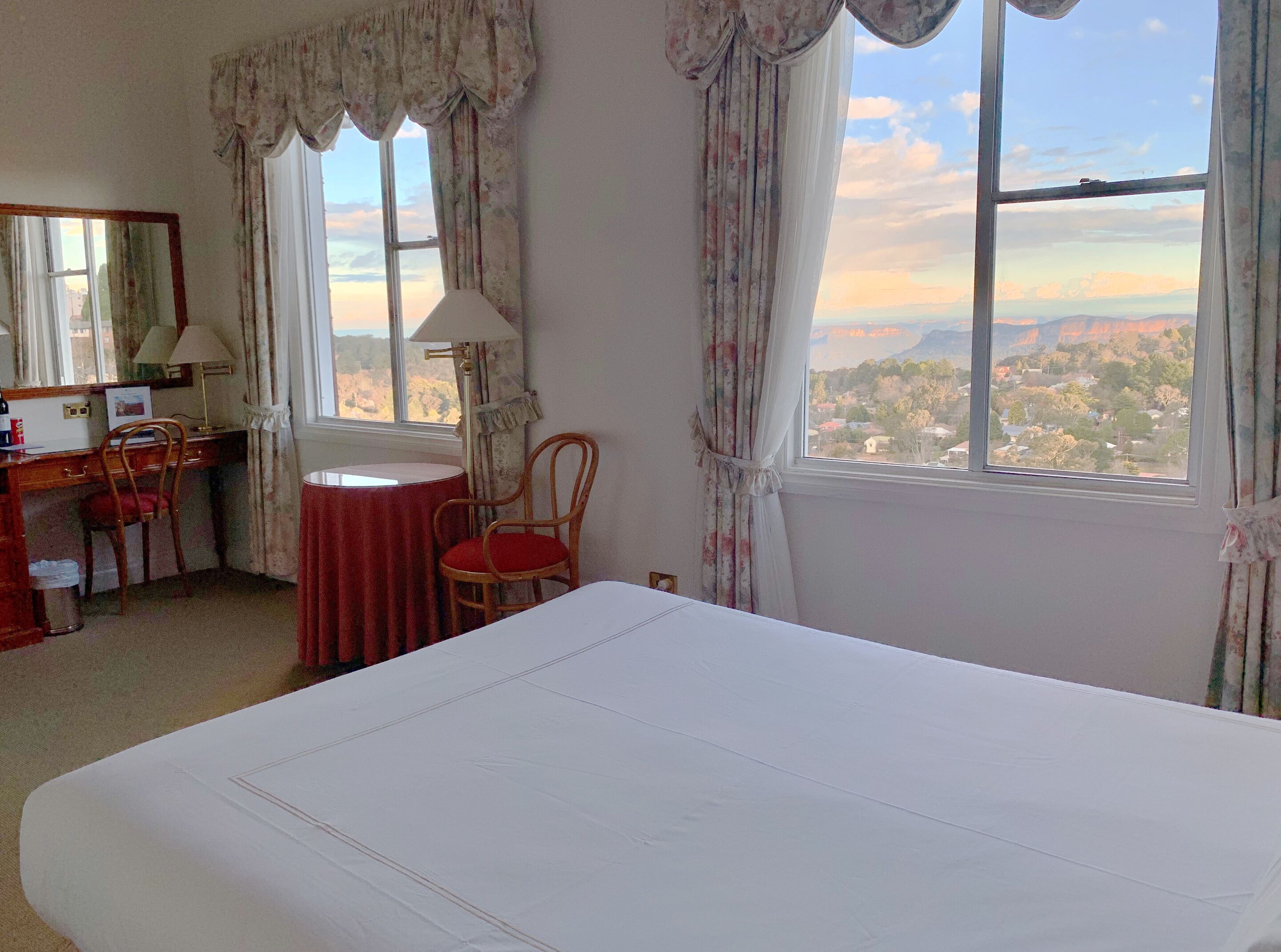 A hotel room with a bed, a desk, two chairs, a lamp, and a view of a landscape through the window.
