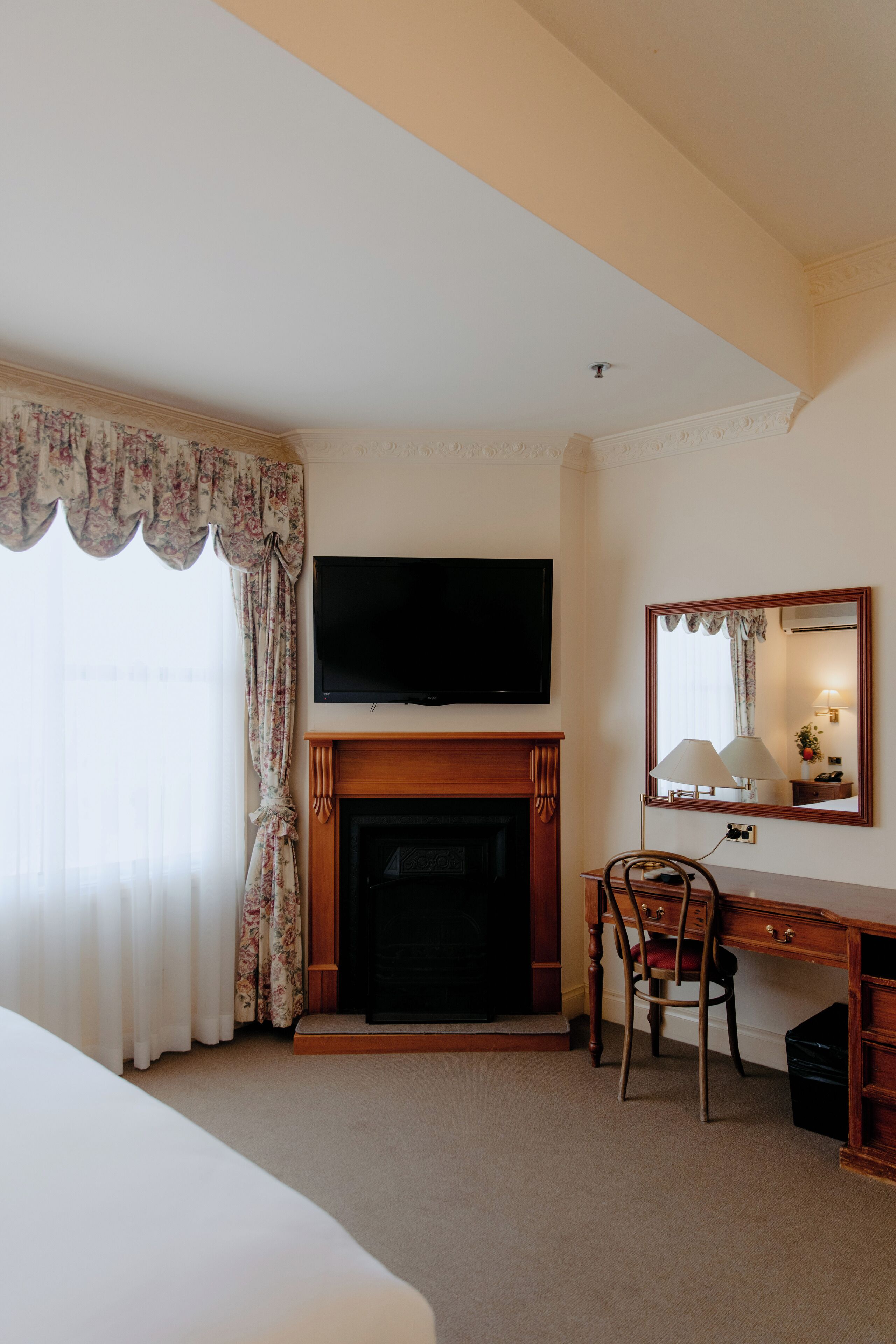 A hotel room with a fireplace, a television mounted above it, a desk with a chair, and a large mirror.