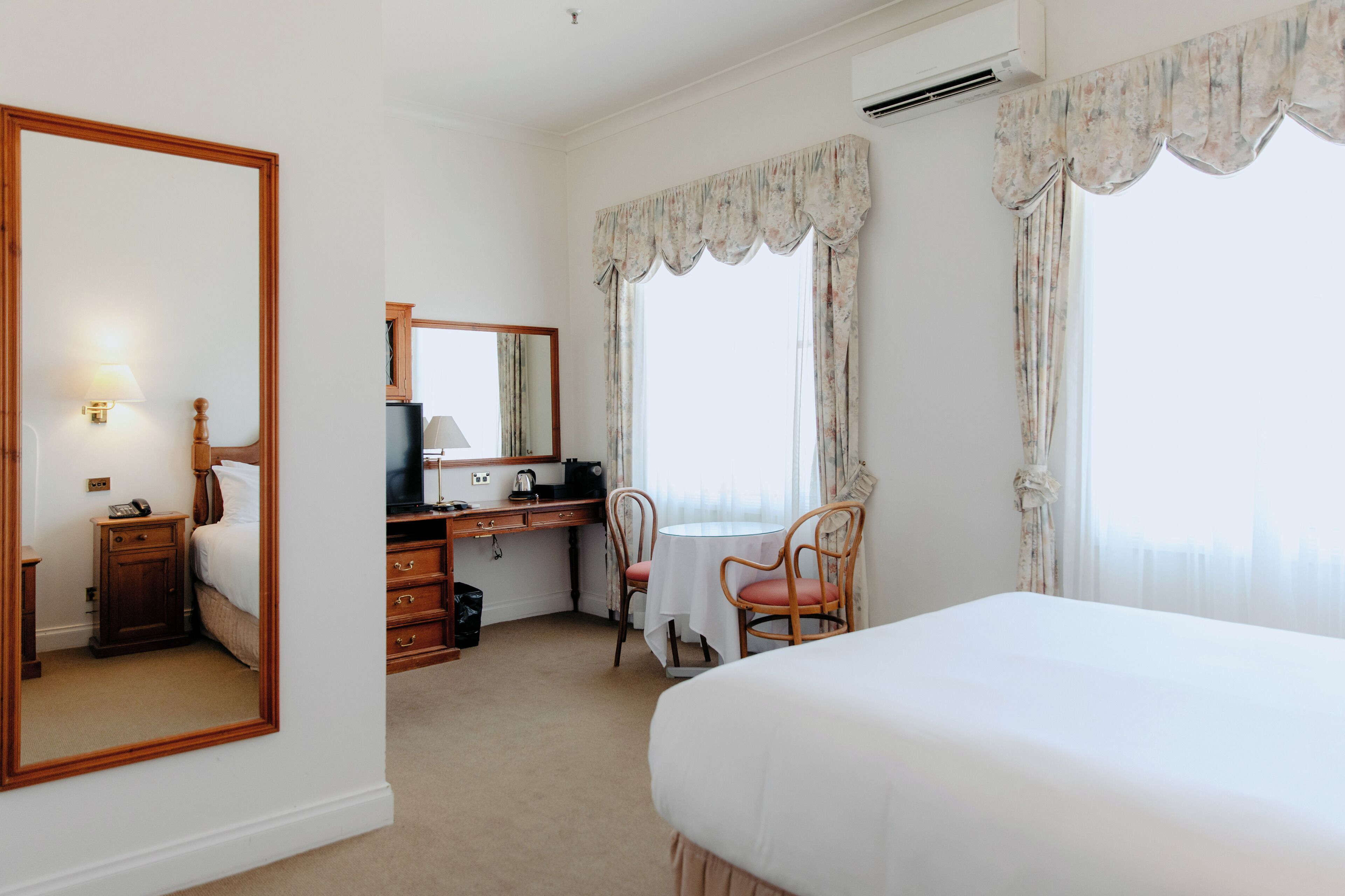 Une chambre d’hôtel comprenant un lit, un bureau avec une chaise, une petite table avec une chaise, un miroir et une fenêtre avec des rideaux.