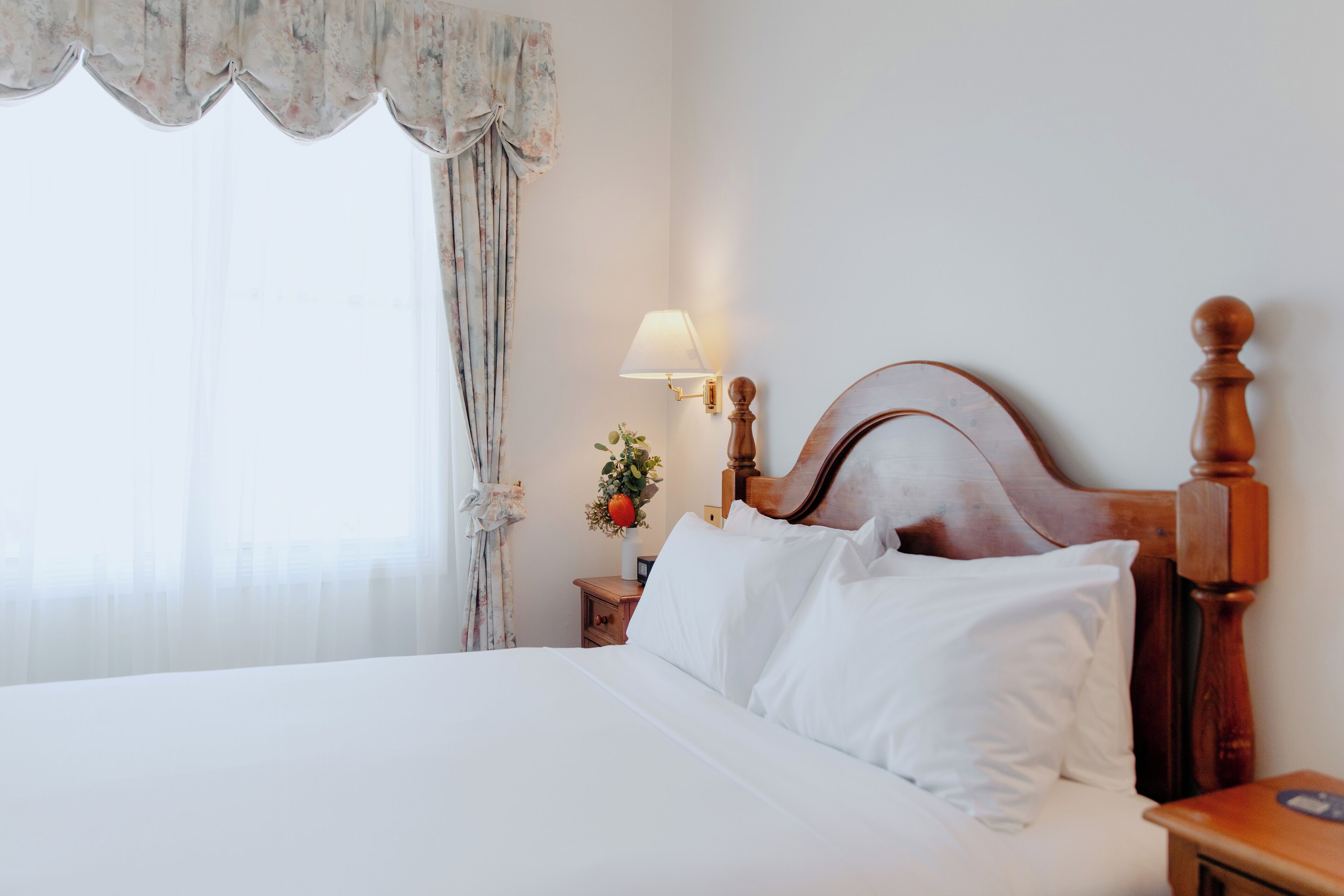 A neatly made bed with white linens, a wooden headboard, a bedside lamp, and a nightstand with a vase of flowers.