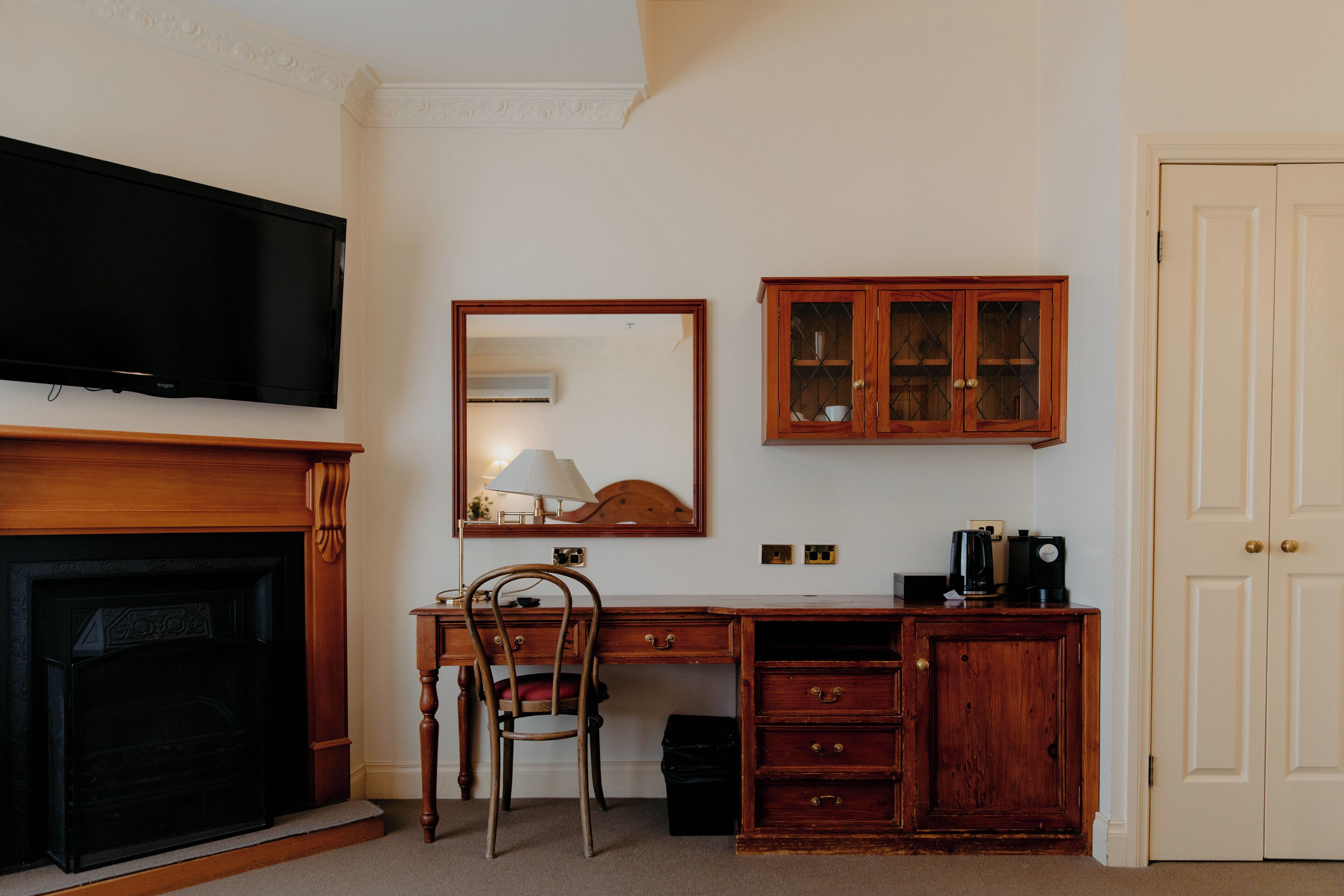 A room with a fireplace, a television mounted above it, a wooden desk with drawers, a chair, a cabinet with glass doors, and a door.