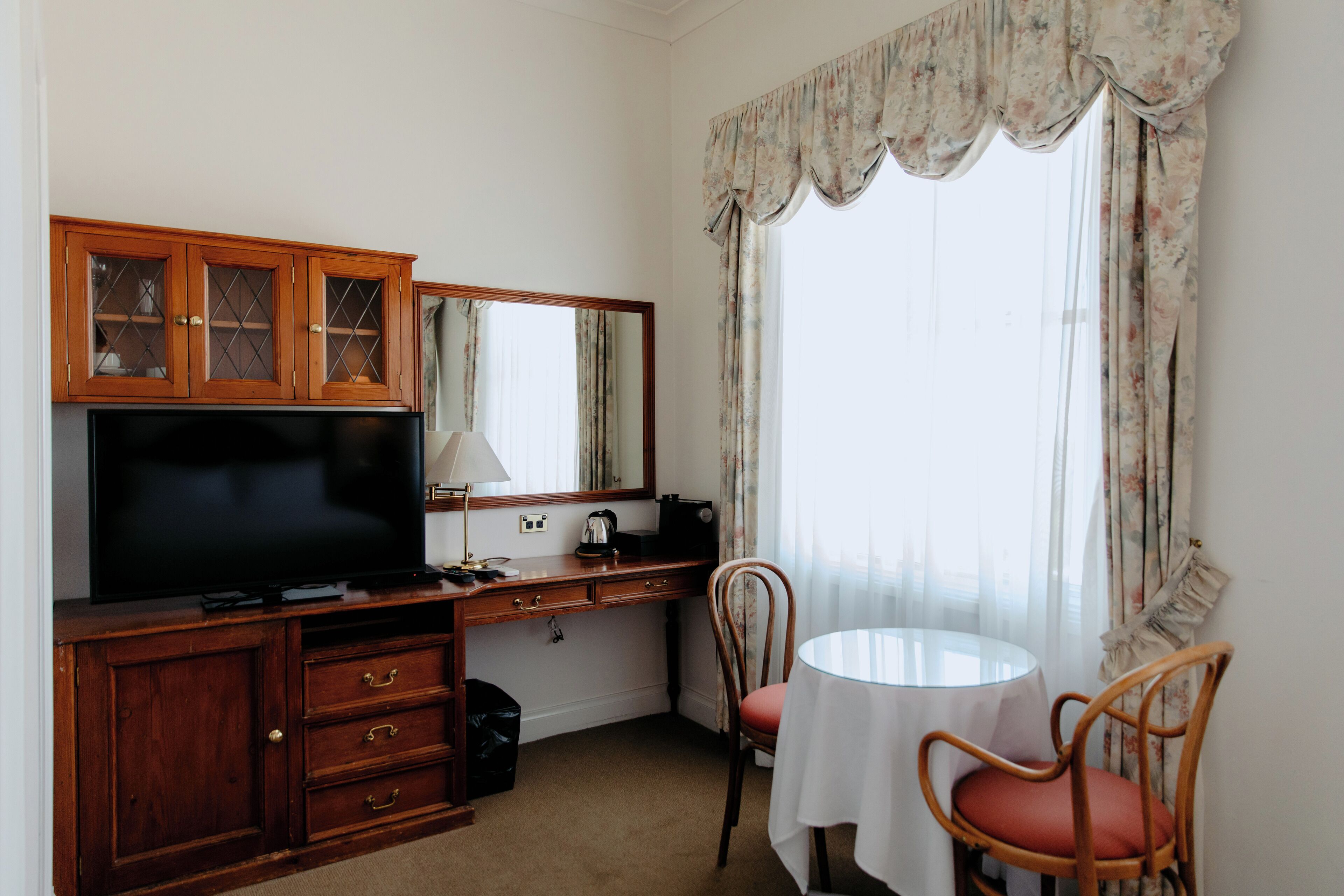 Habitación de hotel con escritorio de madera, televisor de pantalla plana, espejo, pequeña mesa con mantel blanco y dos sillas de mimbre.