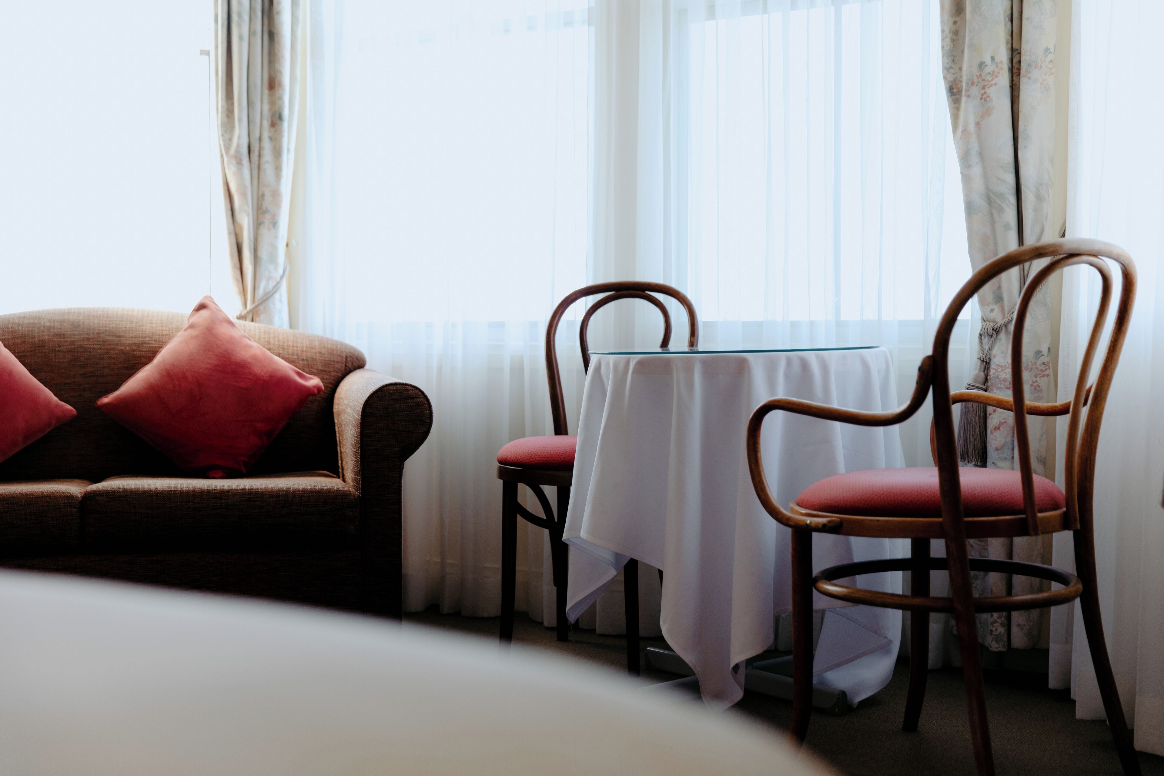 A hotel room with a bed, a brown sofa with red cushions, a wooden chair, and a small table with a white tablecloth.