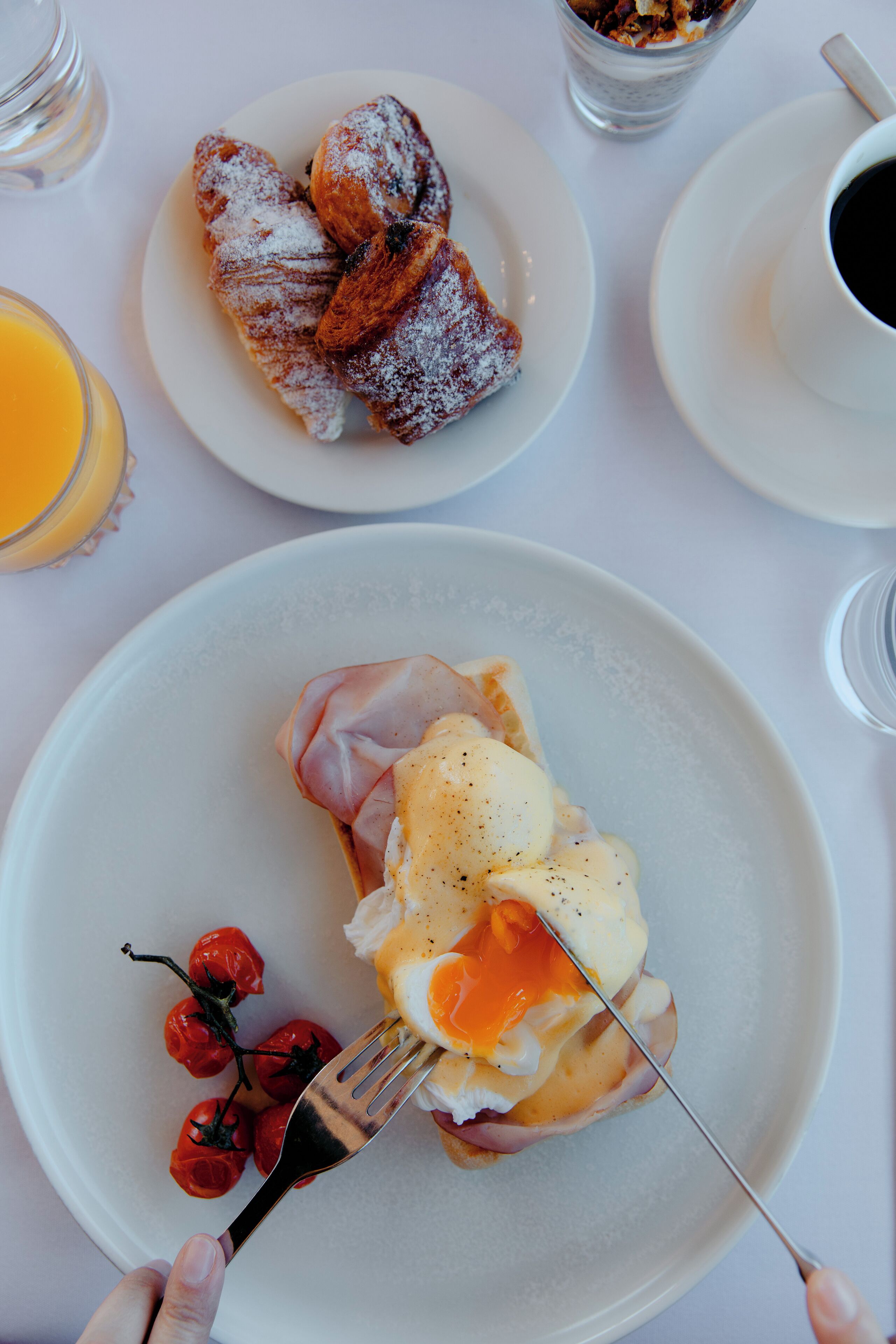 A breakfast plate with ham, eggs, and cherry tomatoes, a plate with pastries, and a cup of coffee on a table.
