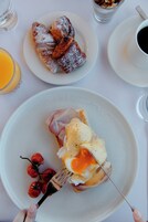 Un plato de desayuno con jamón, huevos y tomates cherry, un plato con pastelería y una taza de café sobre una mesa.