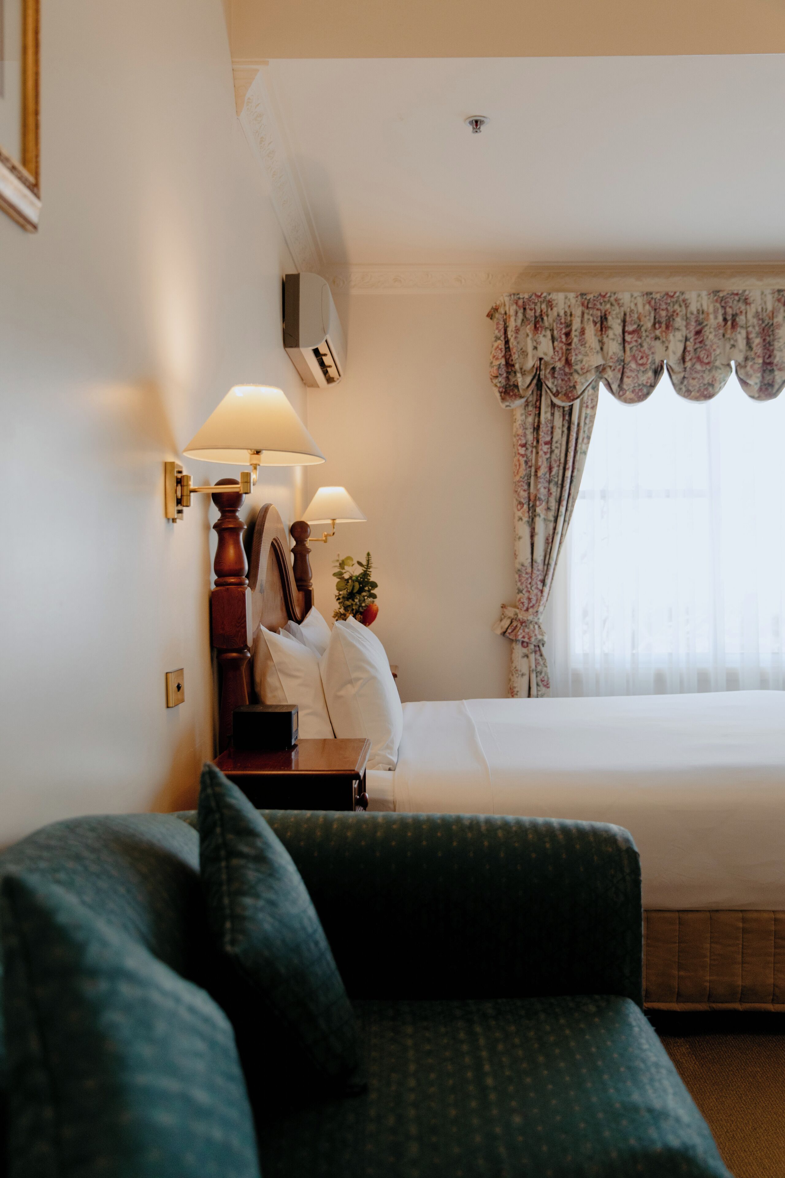 A hotel room with a bed, a nightstand with a lamp, a green armchair, and a window with curtains.
