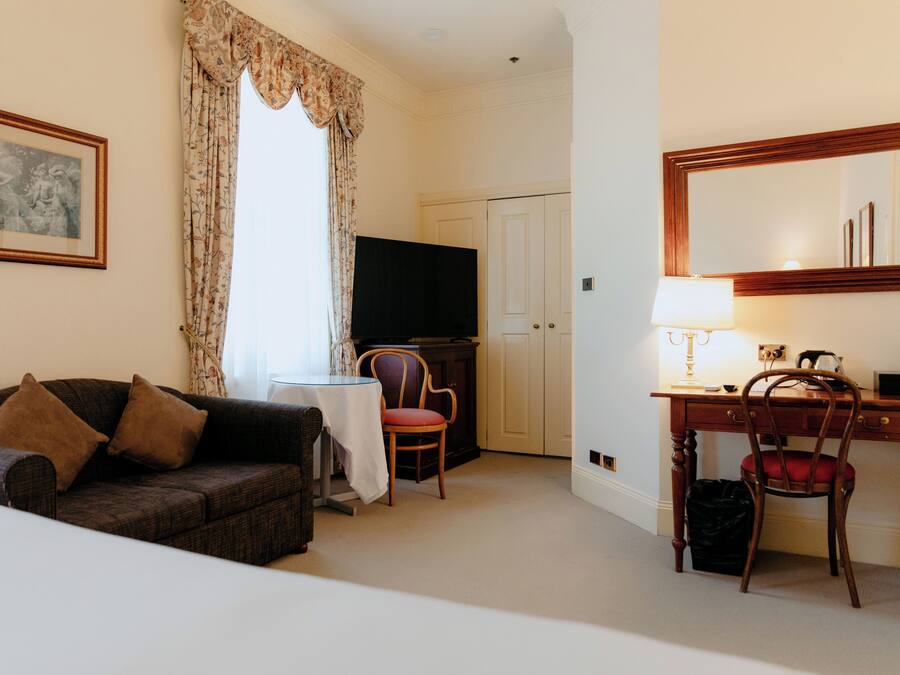 A hotel room with a sofa, a dining table, a chair, a desk with a chair, a television, a lamp, and a mirror.