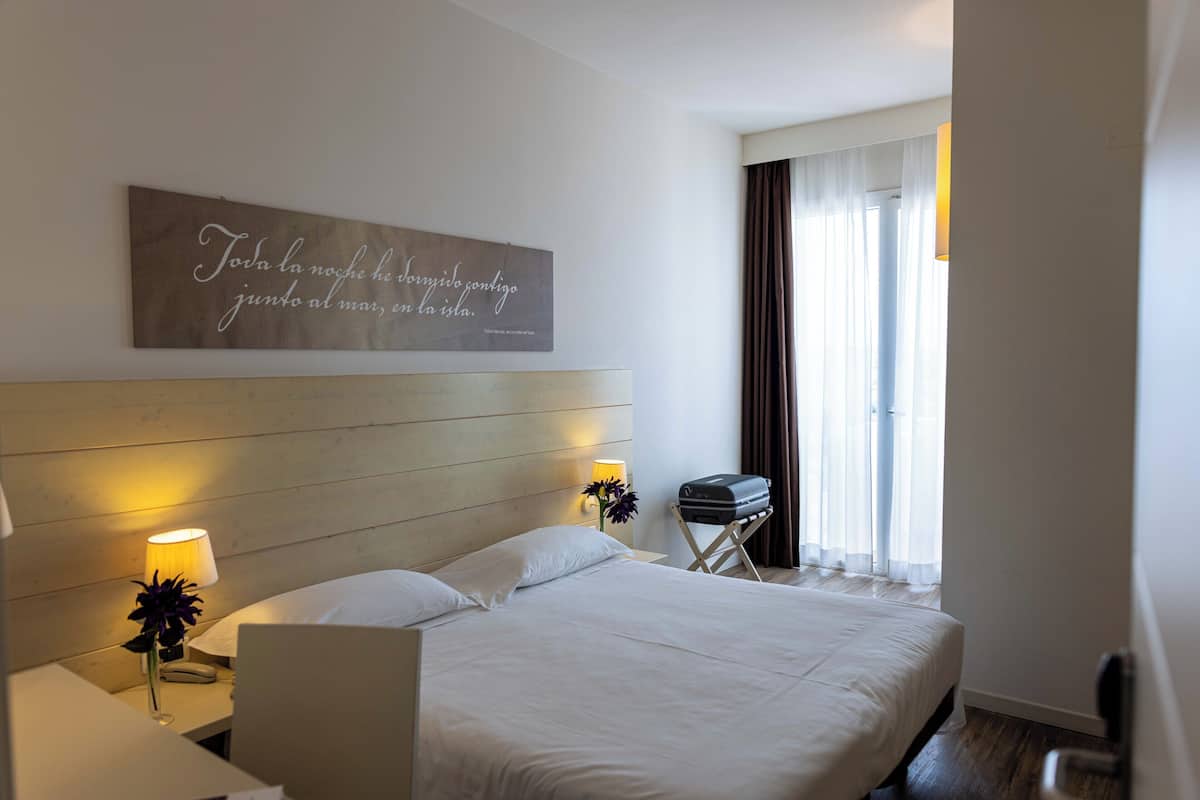 A hotel room with a wooden headboard, a bed with white linens, a bedside lamp, a small table with a lamp and flowers, a suitcase, and a window with curtains.