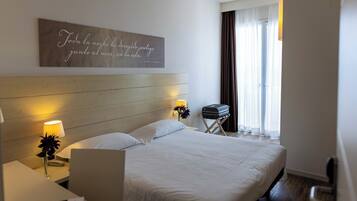 Ein Hotelzimmer mit hölzernem Kopfteil, einem Bett mit weißer Bettwäsche, einer Nachttischlampe, einem kleinen Tisch mit Lampe und Blumen, einem Koffer und einem Fenster mit Vorhängen.