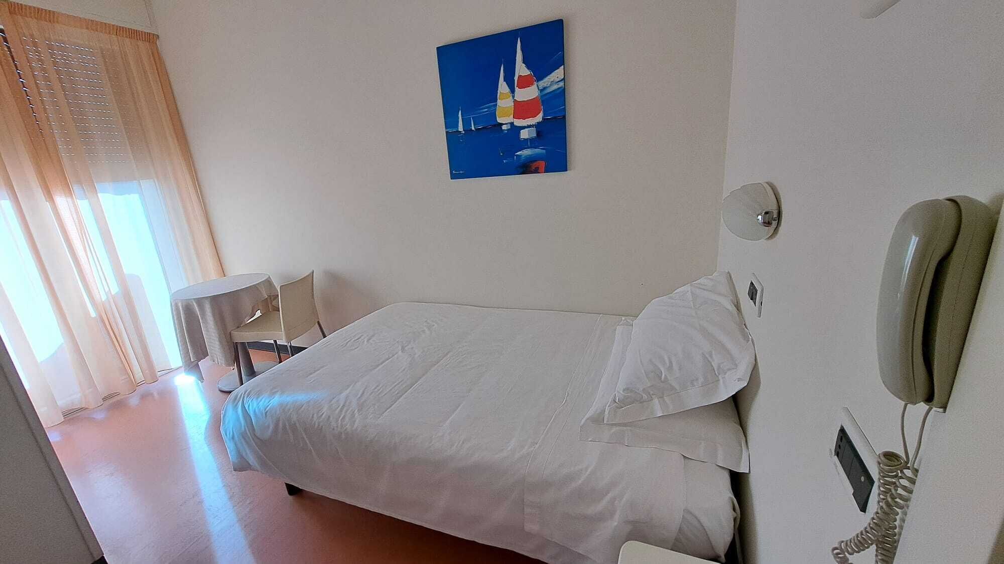 A hotel room with a bed, a bedside phone, a small table with chairs, and a painting of sailboats on the wall.