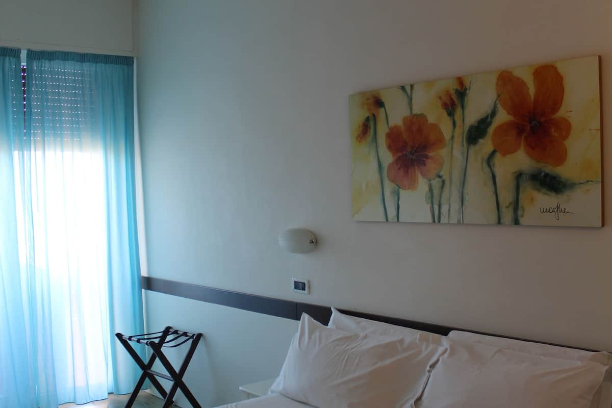 A hotel room with a large bed, a wall-mounted painting of flowers, a window with sheer curtains, and a small table with a chair.