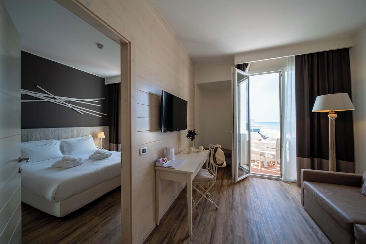 A hotel room with a bed, a desk, a TV, a lamp, and a view of the beach through the door.