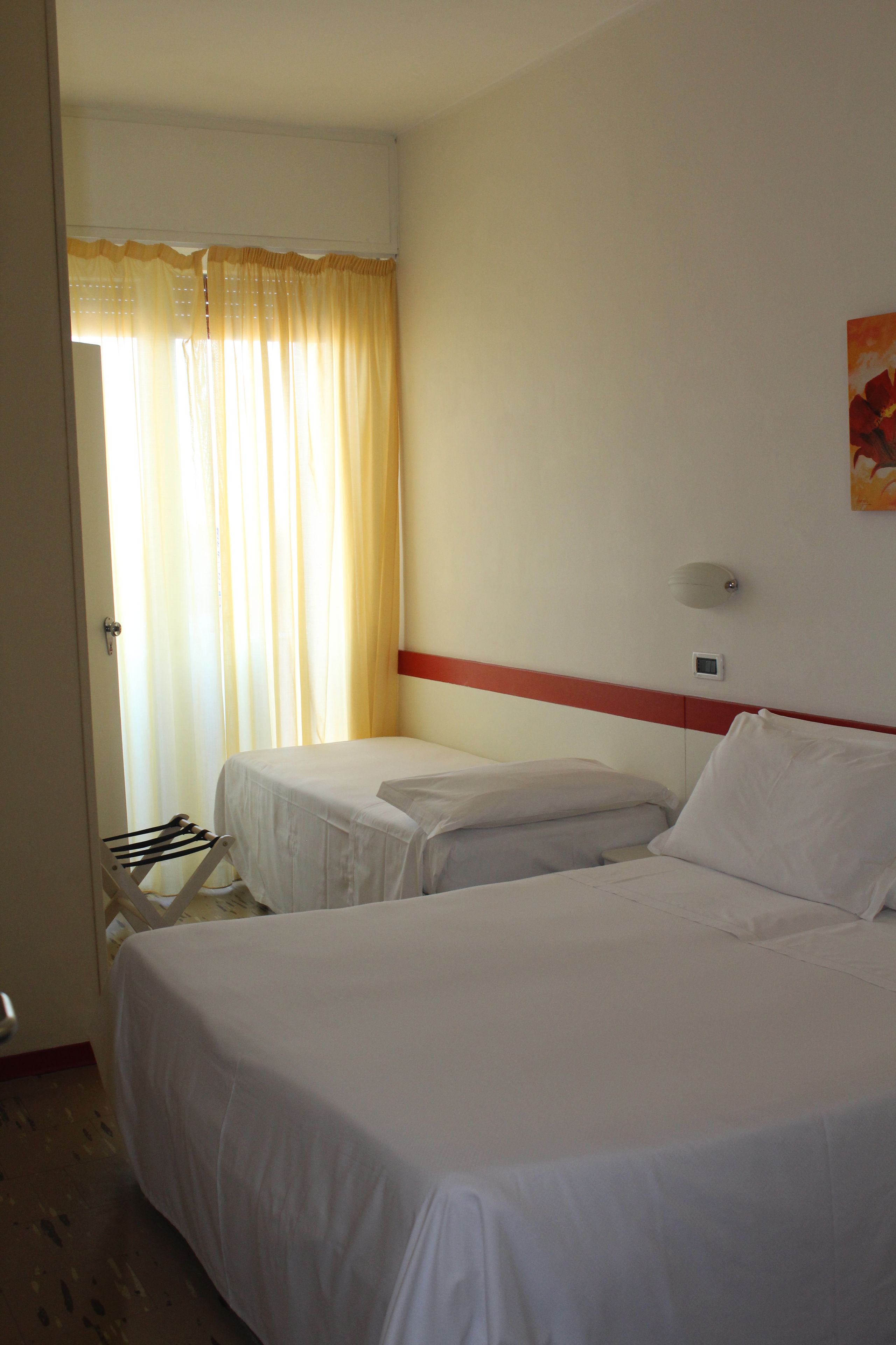 A hotel room with two single beds, a red accent wall, a window with curtains, and a wall-mounted light.