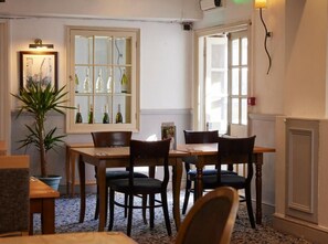 A dining area with a table and chairs, a potted plant, and a window with bottles. - The Sun Hitchin by Greene King Inns (Hitchin)