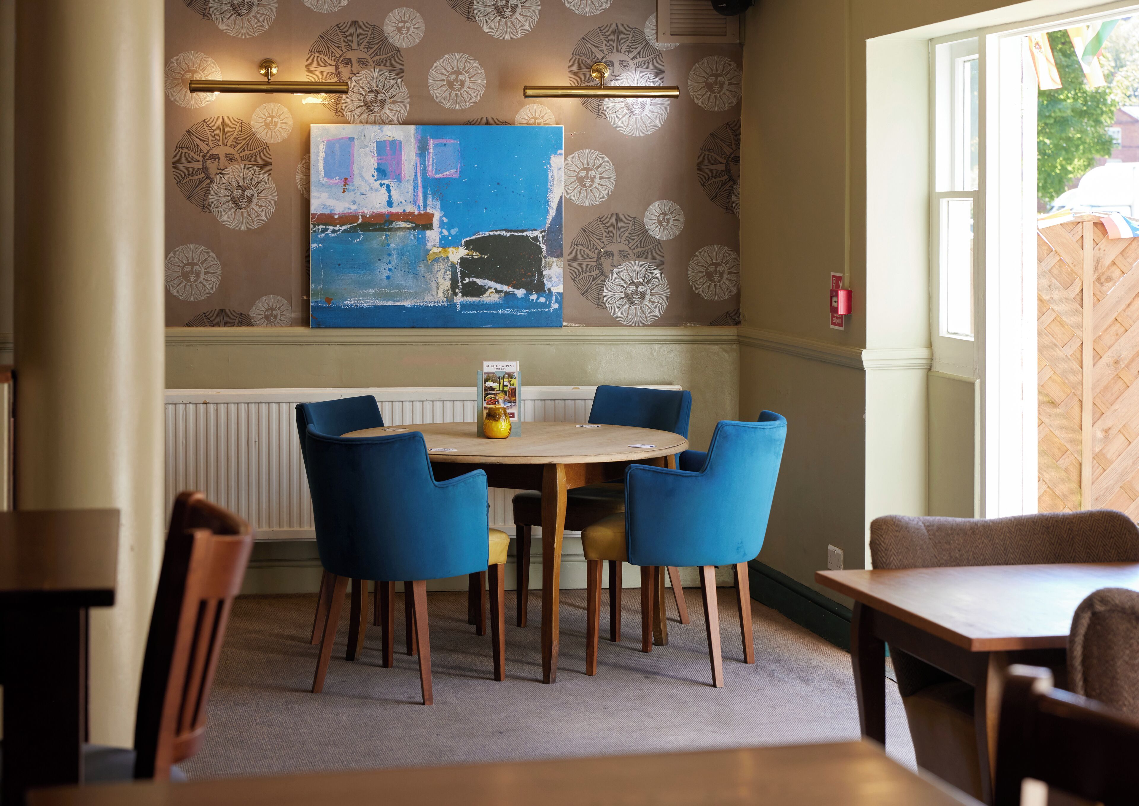 A dining area with a round table, blue chairs, a painting on the wall, and a window with a view of greenery.