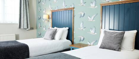 Une chambre d’hôtel avec deux lits, une fenêtre avec des rideaux, un mur orné d’un motif de mouettes et une tête de lit en bois.