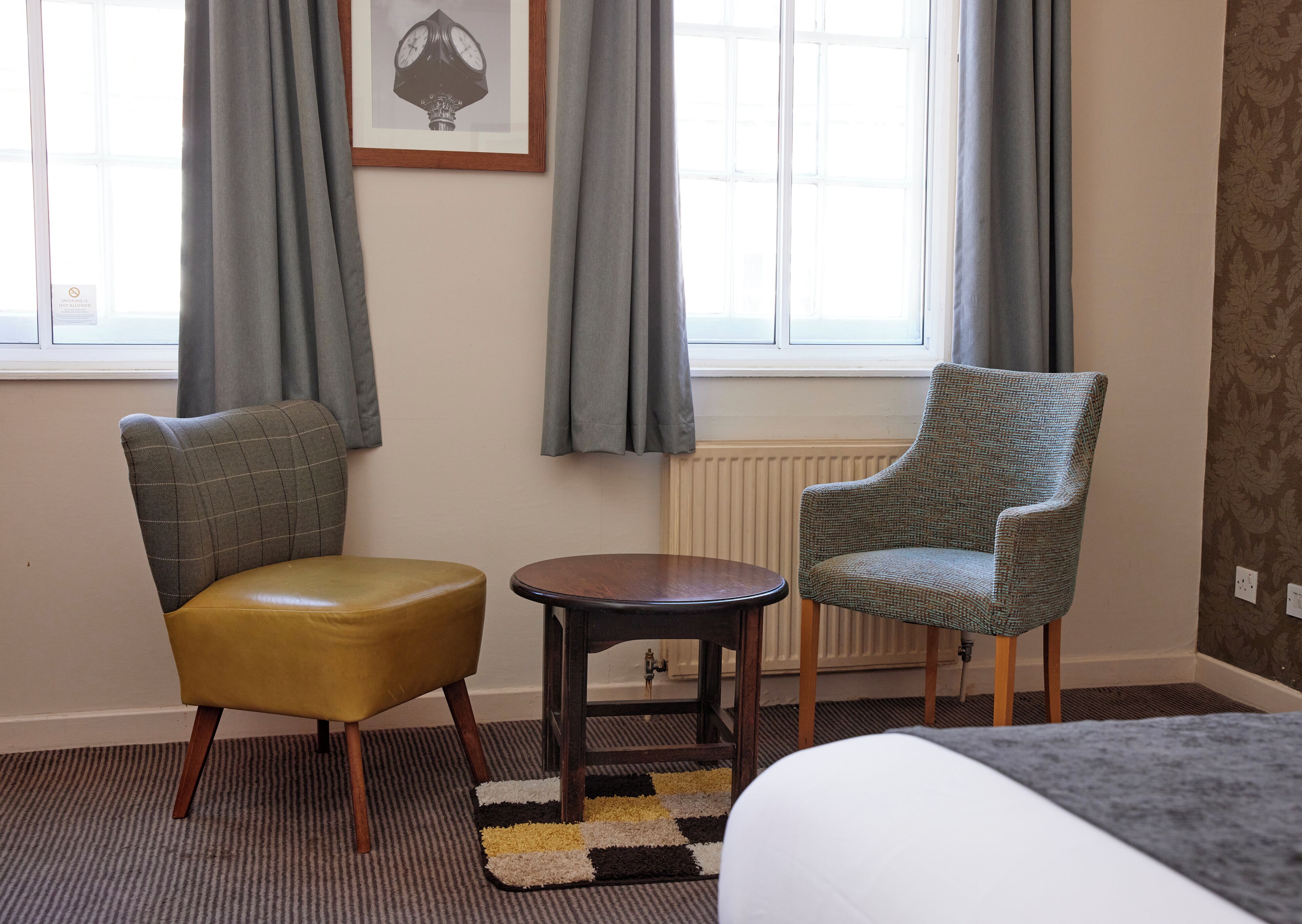 A hotel room with a bed, a small round table, two chairs, a radiator, and a framed picture on the wall.