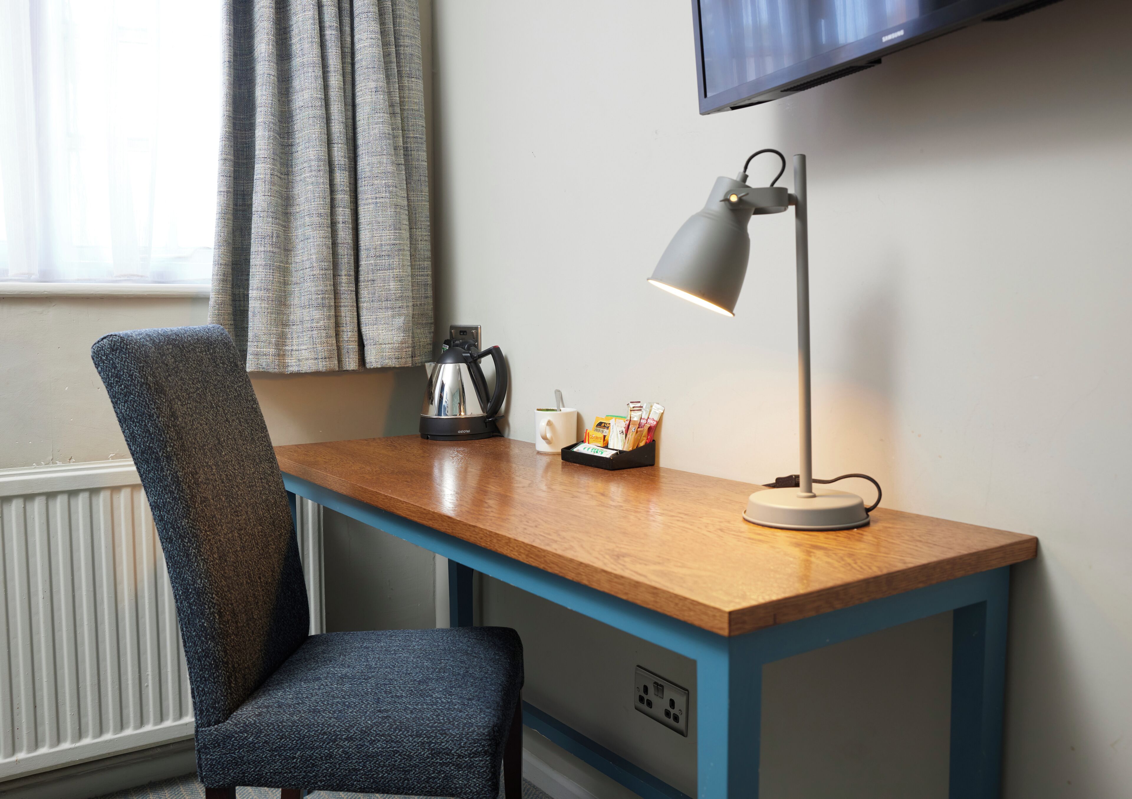 A compact workspace with a wooden desk, a chair, a desk lamp, a kettle, and a flat-screen TV mounted on the wall.