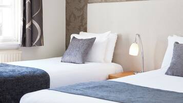 Habitación de hotel con dos camas, cada una con ropa blanca y almohadas grises, una mesita de noche con lámpara y una ventana con cortinas.