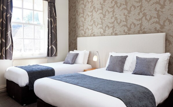A hotel room with two beds, a window with curtains, a lamp, and a patterned wallpaper. - The Sun Hitchin by Greene King Inns (Hitchin)