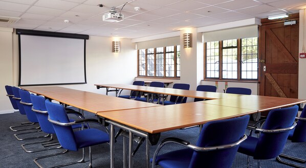 Un salón de conferencias con una larga mesa, sillas azules, una pantalla de proyector y ventanas con persianas.