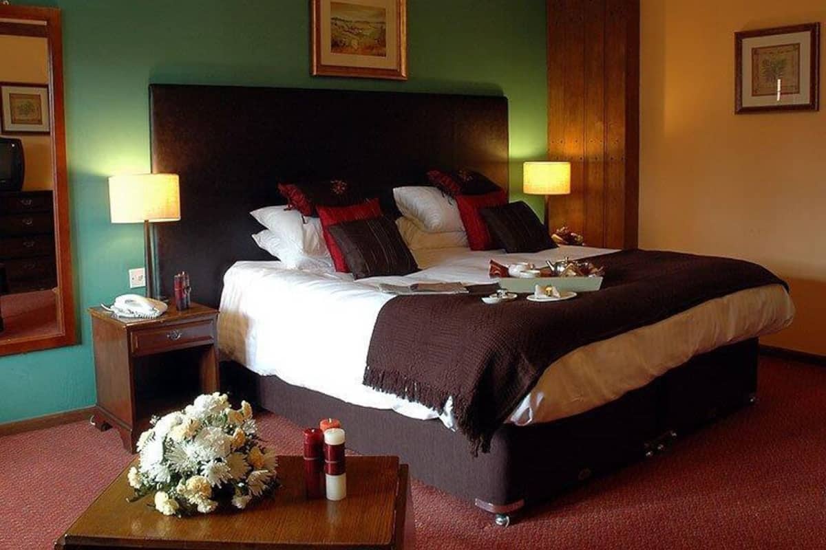 Una habitación de hotel con una cama grande, dos mesitas de noche, una mesita auxiliar, un cabecero de madera y un arreglo floral en el suelo.