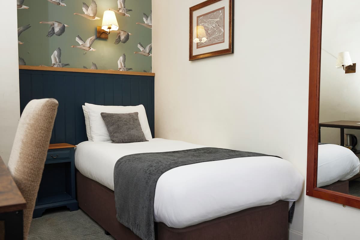 Una cama bien hecha con un edredón blanco y gris, una mesita de noche de madera, una silla, una pared con papel tapiz de pájaros y un cuadro enmarcado.