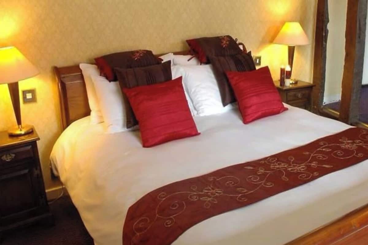 Una cama bien hecha con una colcha roja y varias almohadas, incluidas rojas y marrones. Hay dos lámparas de noche a ambos lados de la cama.